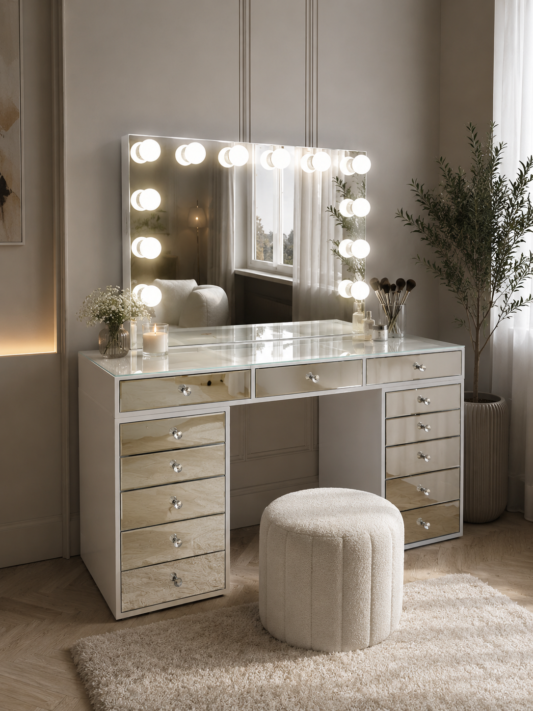 A modern vanity table with a large lighted mirror, mirrored drawers, a clear glass top, and a round tufted ottoman in a softly lit room.