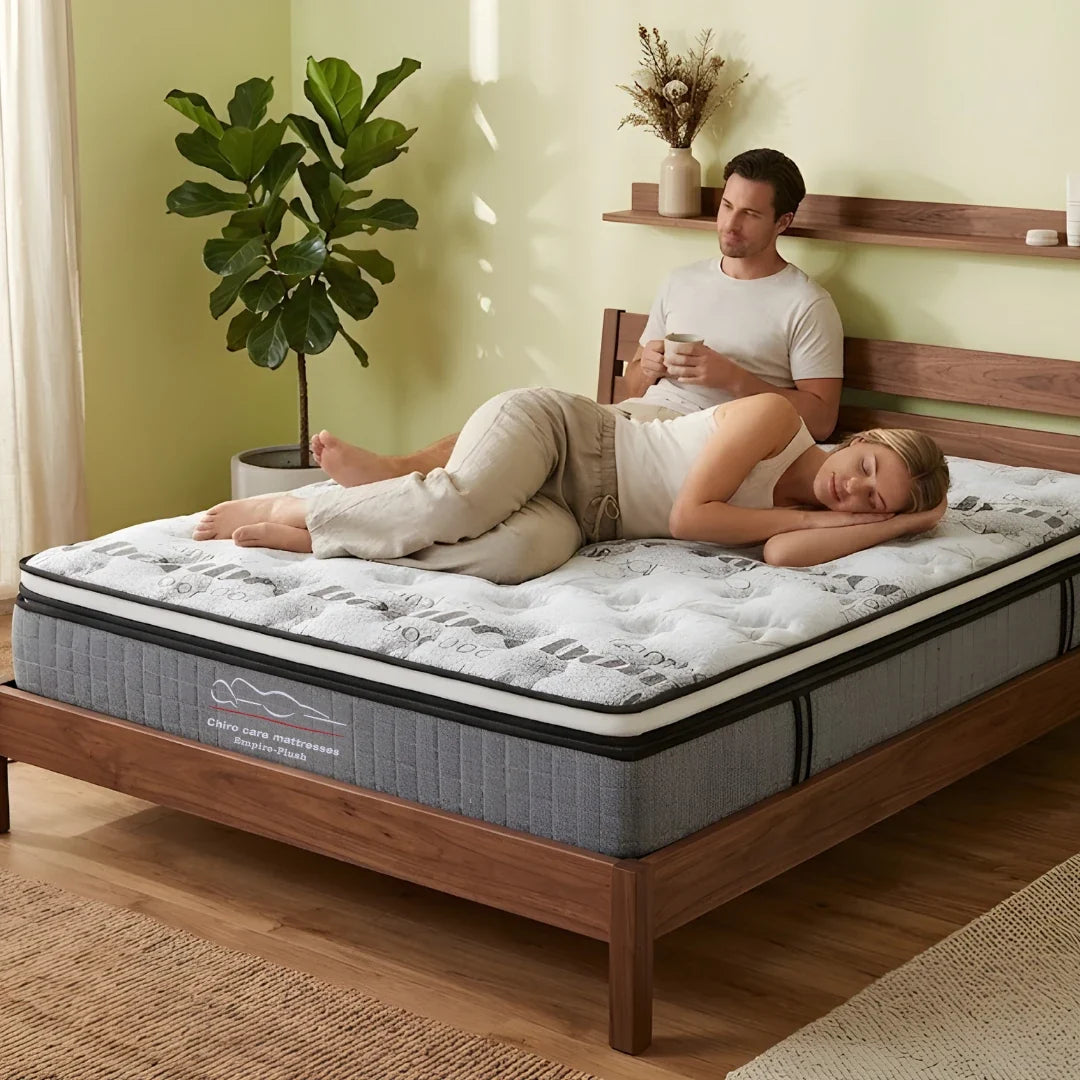 Man and woman lying on a mattress in a bedroom with a plant and shelf in the background.