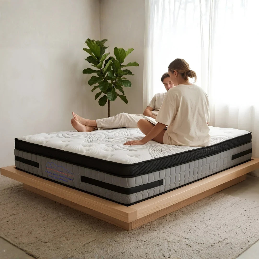 Two people sitting on a mattress in a room with a plant and light curtains.